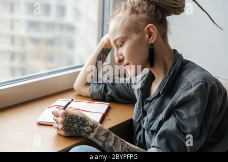 Bild von einem ziemlich konzentrierten jungen Mädchen mit Dreadlocks und Piercing im Innenbereich Notizen in Notizbuch schreiben. Stockfoto