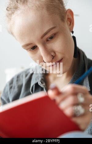 Bild von einem ziemlich konzentrierten jungen Mädchen mit Dreadlocks und Piercing im Innenbereich Notizen in Notizbuch schreiben. Stockfoto