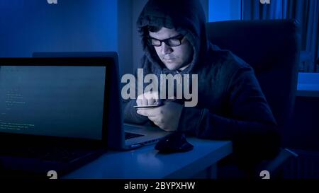Männlicher Hacker in der Haube hält Kreditkarte und arbeitet am Laptop in der Nacht. Cyber-Kriminalität und E-Commerce Stockfoto