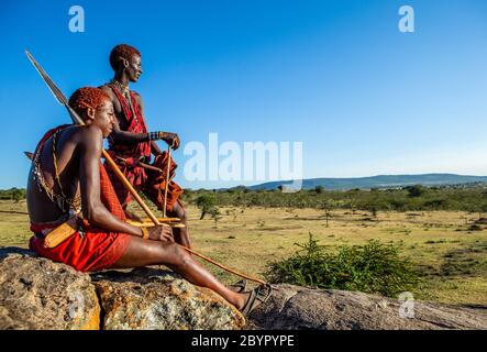 Zwei junge Masai-Krieger sitzen auf einem großen Stein in traditioneller Kleidung mit einem Speer in der Savanne gegen einen blauen Himmel. Tansania, Ostafrika. Stockfoto