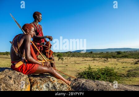 Zwei junge Masai-Krieger sitzen auf einem großen Stein in traditioneller Kleidung mit einem Speer in der Savanne gegen einen blauen Himmel. Tansania, Ostafrika. Stockfoto