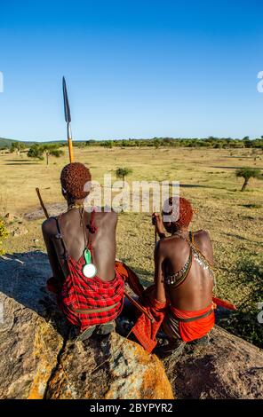 Zwei junge Masai-Krieger sitzen auf einem großen Stein in traditioneller Kleidung mit einem Speer in der Savanne gegen einen blauen Himmel. Tansania, Ostafrika. Stockfoto