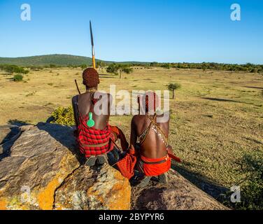 Zwei junge Masai-Krieger sitzen auf einem großen Stein in traditioneller Kleidung mit einem Speer in der Savanne gegen einen blauen Himmel. Tansania, Ostafrika. Stockfoto