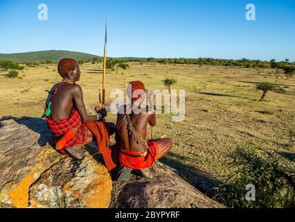 Zwei junge Masai-Krieger sitzen auf einem großen Stein in traditioneller Kleidung mit einem Speer in der Savanne gegen einen blauen Himmel. Tansania, Ostafrika. Stockfoto