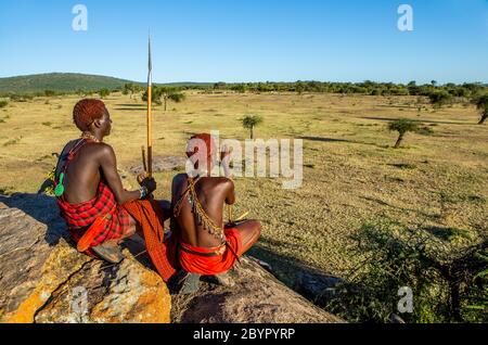 Zwei junge Masai-Krieger sitzen auf einem großen Stein in traditioneller Kleidung mit einem Speer in der Savanne gegen einen blauen Himmel. Tansania, Ostafrika. Stockfoto