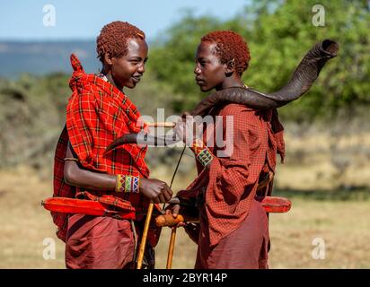 Zwei junge Masai-Krieger in traditioneller Kleidung stehen in der Savanne und sprechen miteinander. Man hält ein rituelles Horn. Tansania, Ostafrika. Stockfoto