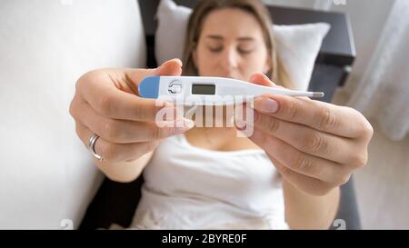 Junge kranke Frau, die auf dem Sofa liegt und ein digitales Thermometer in der Kamera zeigt Stockfoto