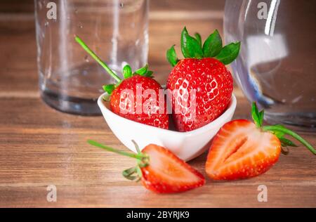 Frische rote Erdbeeren ganz und in Scheiben geschnitten Hälfte und Glas Tasse und Glas Wasser im Hintergrund auf Holz Stockfoto