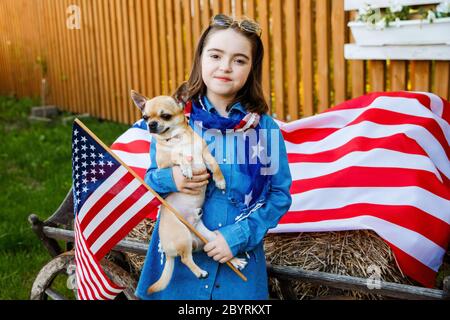 Das Mädchen steht im Hof, in einem Denim-Kleid gekleidet und hält die Flagge der USA und kleinen niedlichen Hund Stockfoto