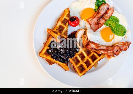 Flacher weißer Teller mit leckerem Frühstück. Das Essen besteht aus belgischen Waffeln, Spiegeleiern, Marmelade, Speck und Basilikum Stockfoto