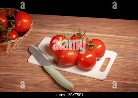 Bouquet von reifen Bio-Tomaten auf einem weißen Schneidebrett und Edelstahl-Messer auf braunem Holztisch und schwarzem Hintergrund Stockfoto