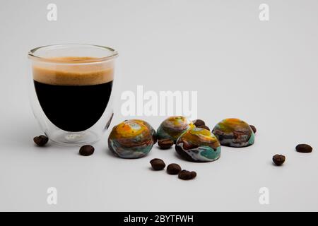 Ein volles, schwarzes, cremiges Espressoglas und bunte, runde Milchschokoladen, die aussehen wie Planeten, die auf weißem Hintergrund isoliert sind, mit Kopierraum, Roa Stockfoto