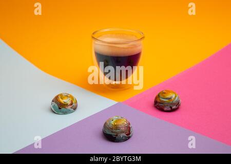 Ein volles, schwarzes, cremiges Espressoglas und bunte, runde Milchschokoladen, die wie Planeten aussehen, die auf einem farbenfrohen Hintergrund mit Kopierraum isoliert sind Stockfoto