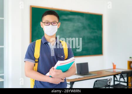 Asiatische Grundschüler Jungen mit Rucksack und Bücher tragen Masken, um den Ausbruch von Covid 19 im Klassenzimmer zu verhindern, während wieder in der Schule öffnen ihre sc Stockfoto