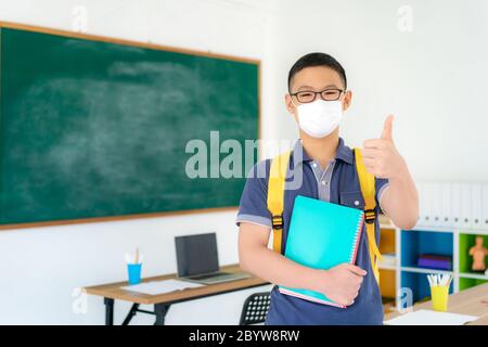 Asiatische Grundschüler Junge Daumen nach oben und trägt Masken, um den Ausbruch von Covid 19 im Klassenzimmer zu verhindern, während wieder in die Schule wieder ihre Schule, Neu Stockfoto