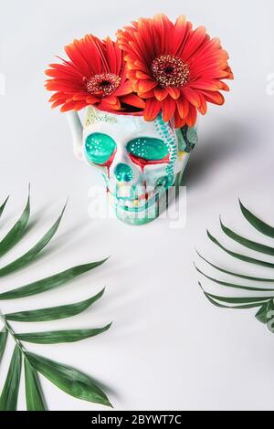 Totenkopf mit korallenfarbenen Gerbera Gänseblümchen und Palmenblättern auf weißem Hintergrund. Kreatives Konzept, diagonale vertikale Komposition. Surreales Gefühl, unwirklich Stockfoto