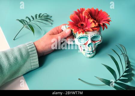 Hand haltenden Becher in Form eines Totenkopfes mit korallenfarbenen Gerbera Gänseblümchen und Palmblättern. Kreatives Konzept auf Kachel grünen Hintergrund, Dia de los Muertos Stockfoto