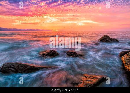 Ein Sonnenuntergang am Meer mit einem lebendigen, farbenfrohen Himmel und Vögeln, die in der Ferne fliegen Stockfoto