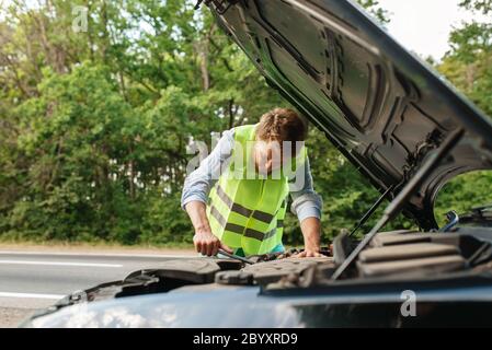 Junger Mann in der reflektierenden Weste an der geöffneten Kapuze, Autoausfall. Gebrochenes Automobil oder Reparatur von Fahrzeug, Probleme mit Auto-Motor auf der Autobahn Stockfoto