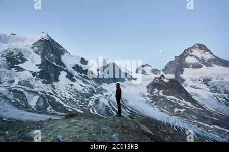 Seitenansicht von allein Touristen genießen wunderschöne Berglandschaft, Mond am Abendhimmel. Trekking, Bergwandern, Mann erreicht Gipfel. Wilde Natur mit herrlichem Ausblick. Sporttourismus in der Schweiz. Stockfoto