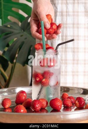 Frau Hand bereitet Erdbeer kalt Getränk mit Eis in Glas auf weißem Hintergrund mit tropischen Blättern. Selektiver Fokus. Stockfoto