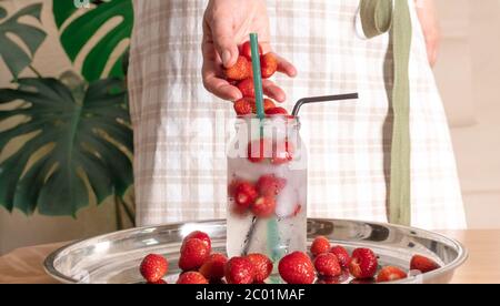 Frau Hand bereitet Erdbeer kalt Getränk mit Eis in Glas auf weißem Hintergrund mit tropischen Blättern. Selektiver Fokus. Stockfoto