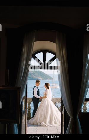 Die Braut und der Bräutigam stehen auf dem Balkon des Hotels mit Blick aufs Meer, ein Blick durch das offene alte Fenster. Fine-Art Hochzeitsfoto in Stockfoto