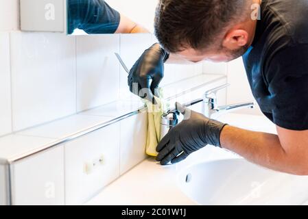 Ein Mann, der ein Waschbecken im Badezimmer mit einem Schaber und einem Mikrofasertuch reinigt, um Kalk und Flecken zu entfernen Stockfoto