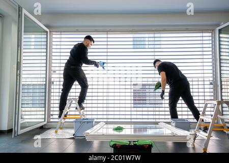 Horizontale Ansicht von zwei männlichen professionellen Reinigern Reinigung Jalousien auf einer großen Fensterfront mit Mikrofasertüchern Stockfoto