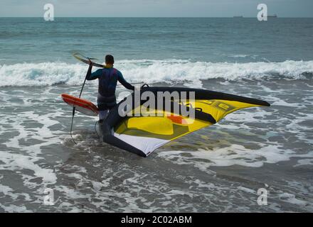 Ein Mann mit einem Neoprenanzug betritt das Meer mit einem handgehaltenen Flügel und einem Tragflügelbrett aus Wingfoil. Stockfoto