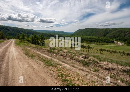 Eine kurvenreiche Straße führt zwischen grünen Bergen mit einem Wald. Unbefestigte Straße. Wolken am blauen Himmel. Sonnig. Horizontal. Stockfoto