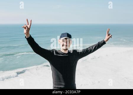 Ein positiver Kerl zeigt seinen Fingern ein Zeichen für Frieden oder Sieg vor dem Hintergrund des Meeres oder Ozeans. Stockfoto