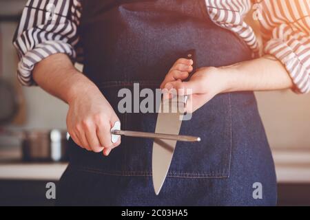 Ein Koch in einem gestreiften Hemd und blauer Schürze steht in der Küche und schärft die Klinge eines Stahlmessers im Sonnenlicht. Stockfoto