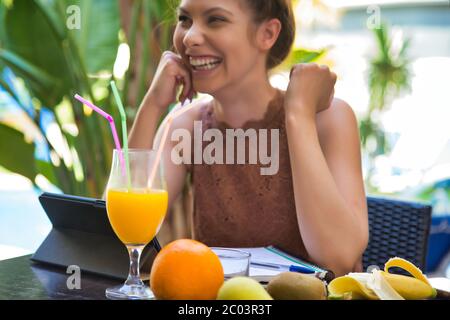 Selektive Konzentration auf einen Fruchtsaft auf einem Terrassentisch mit mehreren Obststücken und einem lächelnden Mädchen im Hintergrund vor einer Tablette und einem Notizblöcken Stockfoto