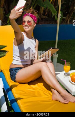 Vertikale Aufnahme einer Frau mit Turban und Sonnenbrille auf einer Sonnenliege mit einer Tablette und einem Teller mit Obst sitzen und ein Selfie in einem Garten machen Stockfoto