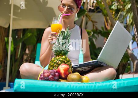 Frau mit Sonnenbrille, die Saft mit Stroh trinkt, während sie auf einer Sonnenliege sitzt und mit einem Laptop in einem Garten mit einem Tisch mit Frühstück arbeitet Stockfoto