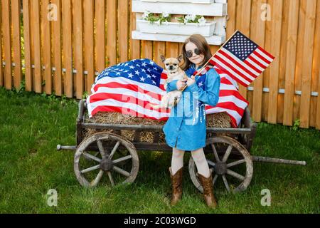 Das Mädchen steht im Hof, in einem Denim-Kleid gekleidet und hält die Flagge der USA und kleinen niedlichen Hund Stockfoto