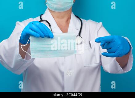 Doktor in einem weißen Mantel und Maske hält einen Stapel von schützenden Einweg-Gesichtsmasken, Arzt steht auf einem blauen Hintergrund Stockfoto