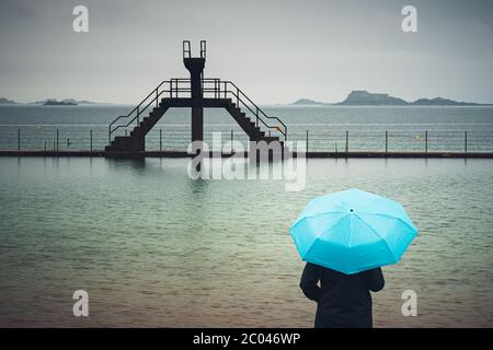 Frau mit blauem Schirm, die an einem launischen und regnerischen Tag vor dem Meerwasser-Schwimmbad Bon Secours steht, St Malo, Frankreich. Stockfoto