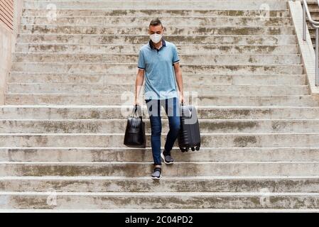 Ein schöner junger Mann auf der Straße, in lässiger Kleidung, trägt eine Gesichtsmaske, trägt eine schwarze Ledertasche in der einen Hand und einen grauen Koffer in der ot Stockfoto