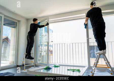 Horizontale Ansicht von zwei männlichen professionellen Reinigern Reinigung Jalousien auf einer großen Fensterfront mit Mikrofasertüchern Stockfoto