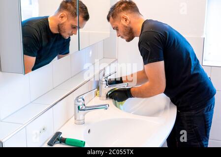 Ein Mann, der ein Waschbecken im Badezimmer mit einem Schaber und einem Mikrofasertuch reinigt, um Kalk und Flecken zu entfernen Stockfoto