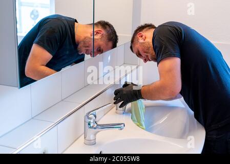 Ein Mann, der ein Waschbecken im Badezimmer mit einem Schaber und einem Mikrofasertuch reinigt, um Kalk und Flecken zu entfernen Stockfoto