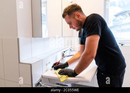 Ein Mann, der ein Waschbecken im Badezimmer mit einem Schaber und einem Mikrofasertuch reinigt, um Kalk und Flecken zu entfernen Stockfoto