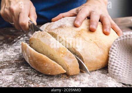 Detail der Hände Schneiden eines Laib rustikalen Brotes in Scheiben auf einem Holztisch voller Mehl, Konzept der gesunden Ernährung zu Hause Stockfoto