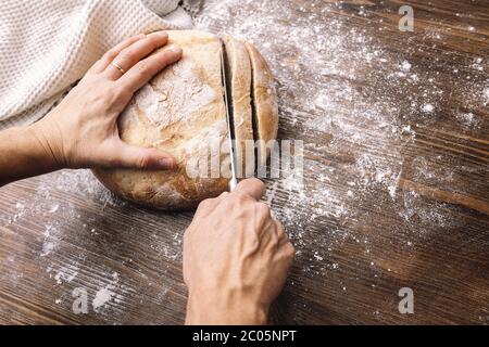 Hände Schneiden ein Laib rustikales Brot in Scheiben auf einem Holztisch voller Mehl, Konzept der gesunden Ernährung zu Hause Stockfoto