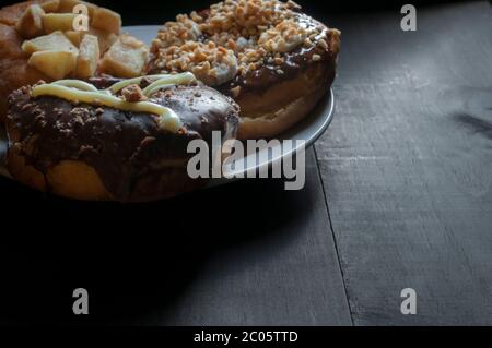 Drei Donuts mit Schokolade auf einem weißen Teller auf einem rustikalen Tisch, der von Fensterlicht, dunkler Lebensmittelfotografie oder dezem Lichtkonzept beleuchtet wird. Stockfoto