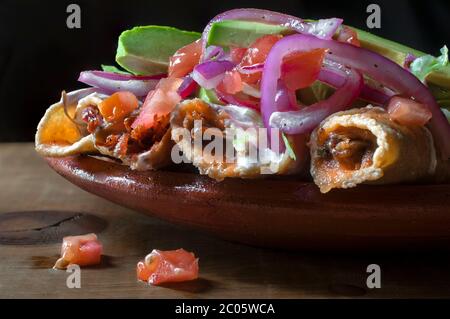 Tacos dorados auf einem mexikanischen Lehm oder Schlammteller, mit lila Zwiebeln, Avocado, Tomatencreme und würziger Salsa. Mexikanisches Essen auf schwarzem Hintergrund. Stockfoto