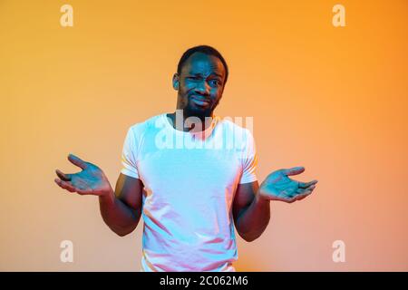 Unsicher, fühlt sich schuldig, wtf. afroamerikanischer Mann modernes Porträt auf gradient orange Studio Hintergrund in Neon. Schönes Afro-Modell. Konzept der menschlichen Emotionen, Gesichtsausdruck, Verkauf, Anzeige. Stockfoto