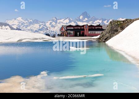 Vorbei am Nufenenpass mit Schnee, Restaurant und kleinem Bergsee, hinter schneebedeckten Gipfeln Finsteraarhorn links und Lauteraarhorn rechts Stockfoto